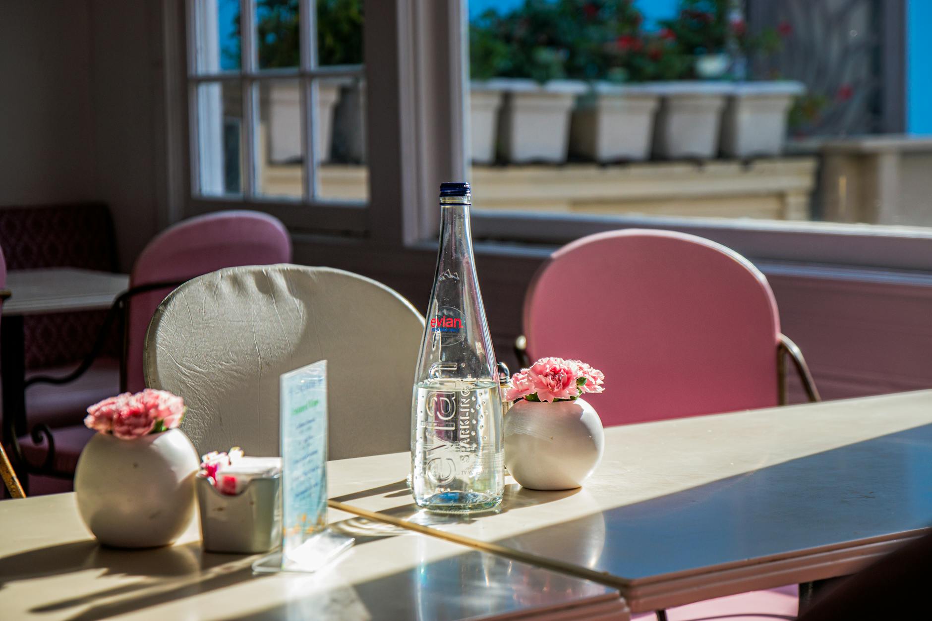 crochet water bottle holder - Sunlight casts shadows on a café table with a water bottle, pink flowers, and chairs.