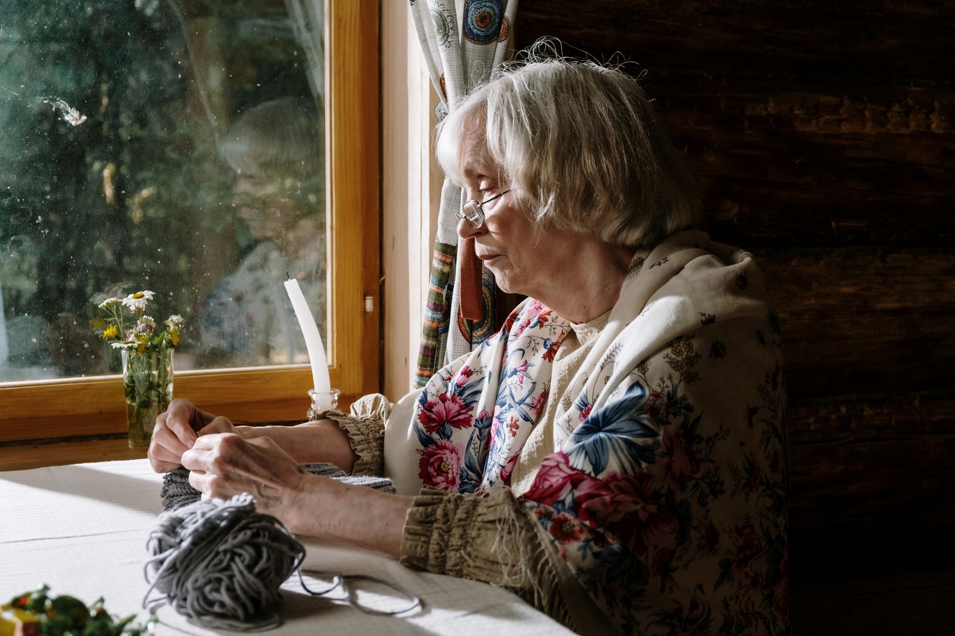 hooded shawl crochet pattern - Senior woman knitting by a sunlit window in cozy interior, embracing a peaceful hobby.