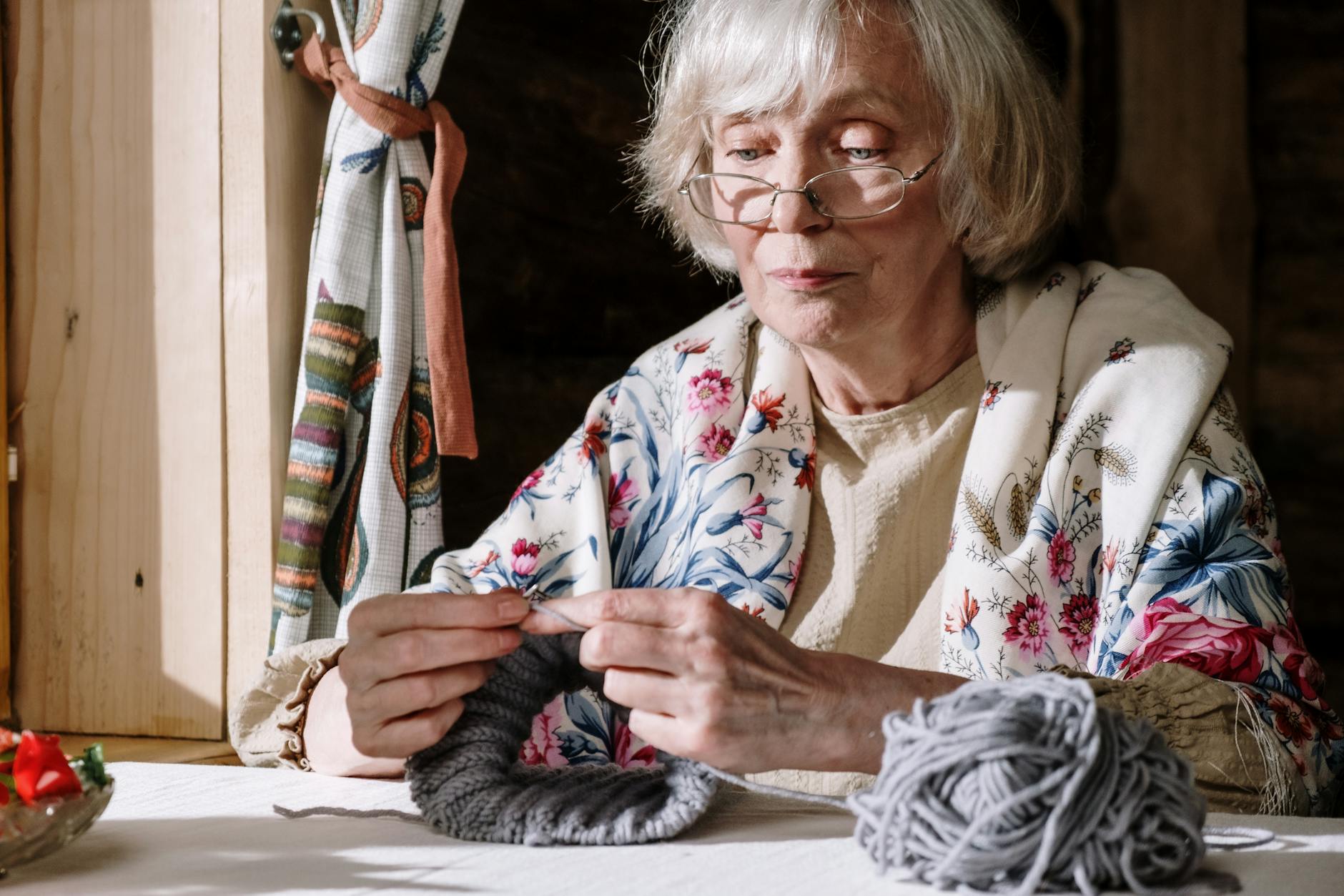 hooded shawl crochet pattern - Senior woman knitting with gray yarn near a sunlit window, showcasing peaceful retirement acti