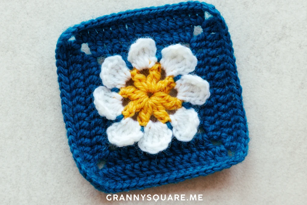 3D flower granny square with yellow bobble stitch center and white petals on blue background