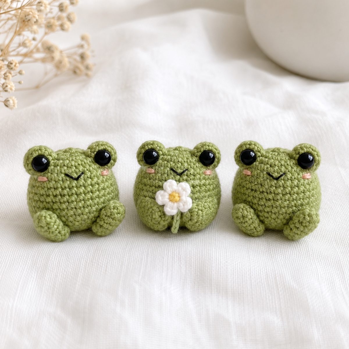 Three tiny green amigurumi crochet frogs with big safety eyes, one holding a small white daisy, on white linen