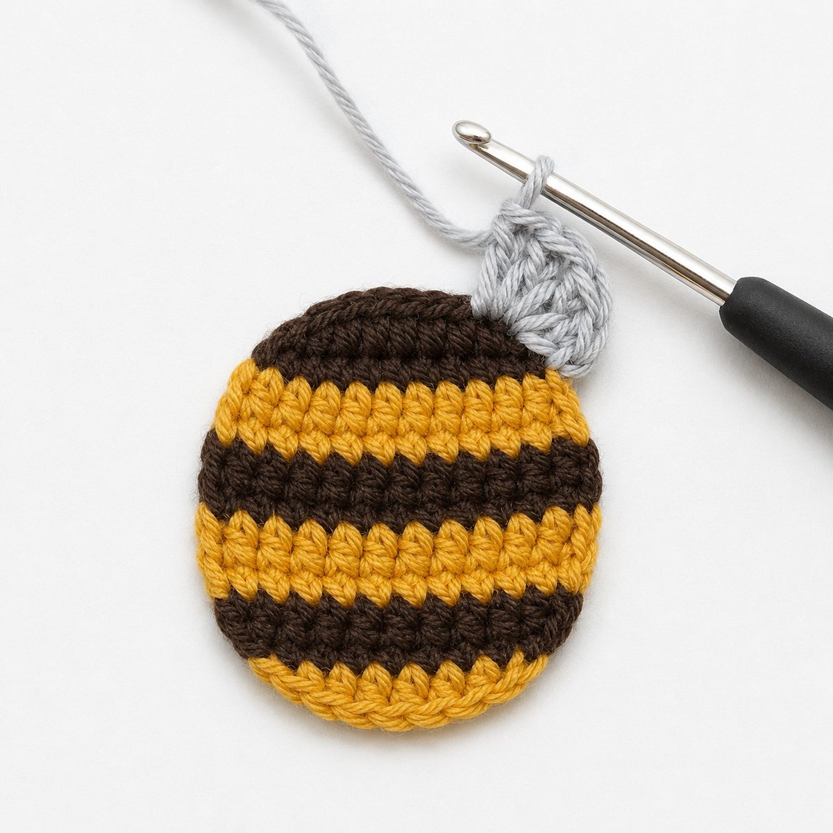 Pale gray wing being crocheted onto the edge of the flat striped bee body, hook holding a loop of gray yarn