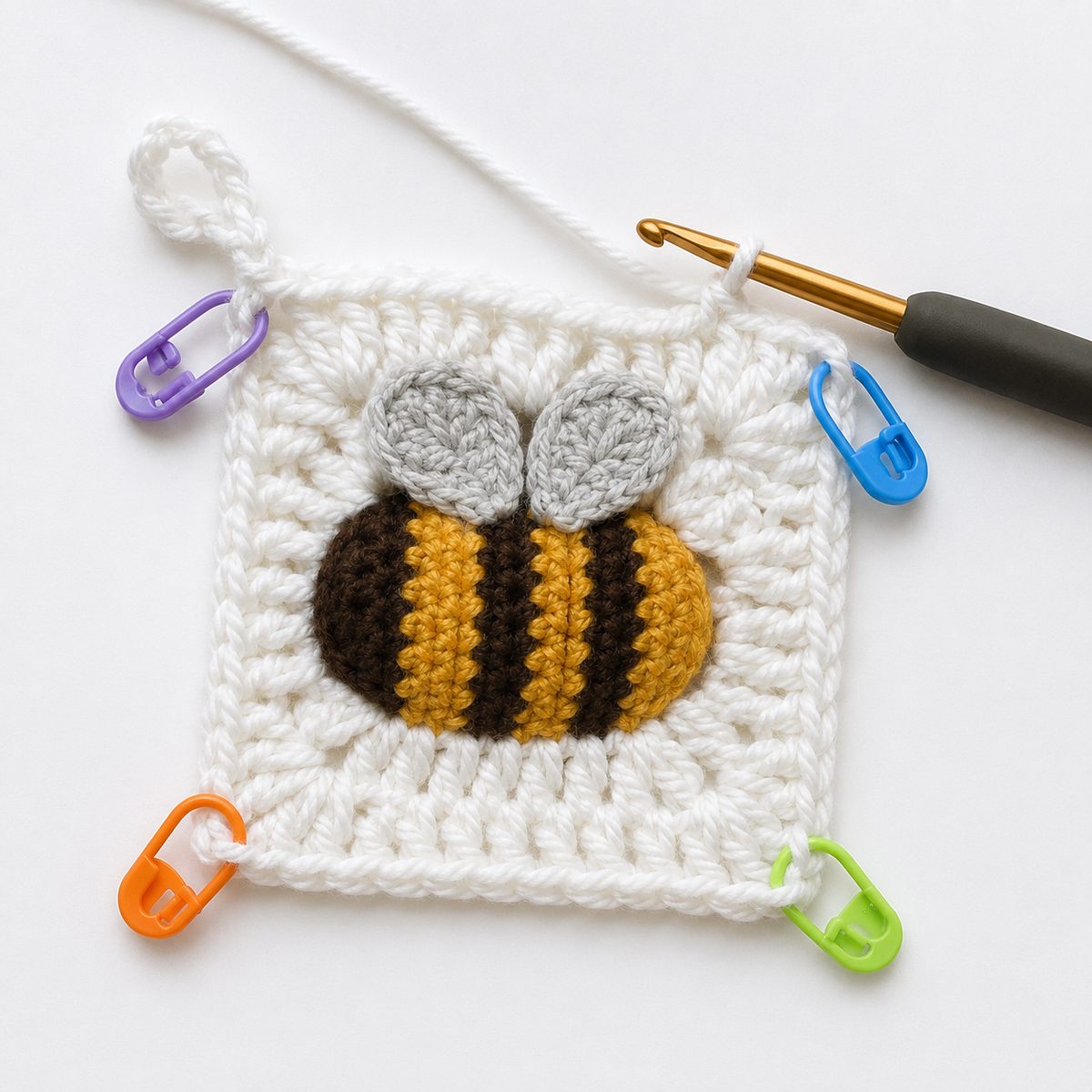 White squaring round worked around the striped bee with four colorful stitch markers at the corner positions