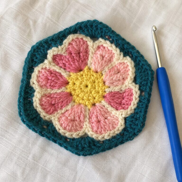 Handmade crochet African flower hexagon motif with pink and cream petals radiating from a yellow center with dark teal border, on white linen