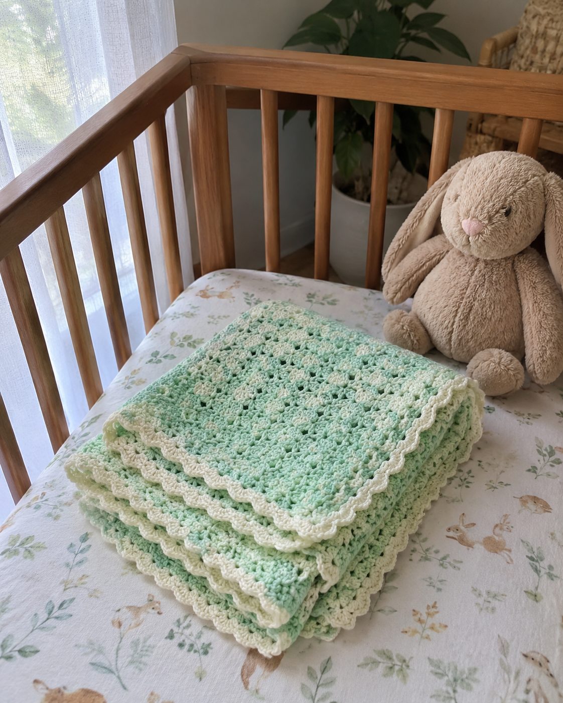 Folded handmade mint green and cream crochet baby blanket with a scalloped shell-stitch edge lying on a wooden crib beside a plush bunny toy in a real nursery