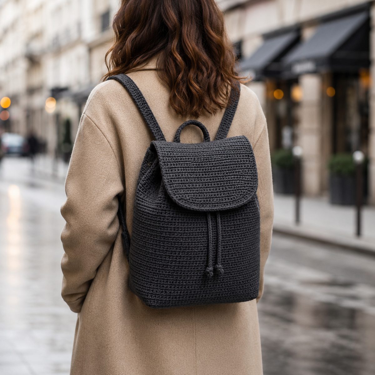 Charcoal grey minimalist crochet backpack with flap worn on the back of a person in a beige coat on a city street