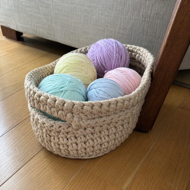 Handmade beige crochet basket in chunky t-shirt yarn holding balls of pastel yarn, on a wooden floor beside a chair leg