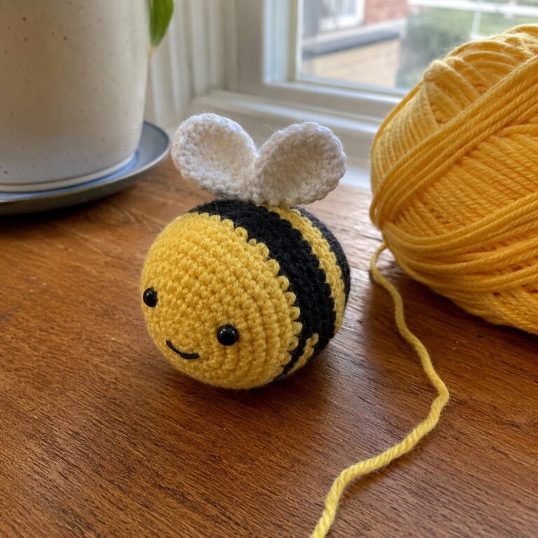 Handmade crochet bee amigurumi with yellow and black striped body and small white wings sitting on a wooden table beside a ball of yellow yarn