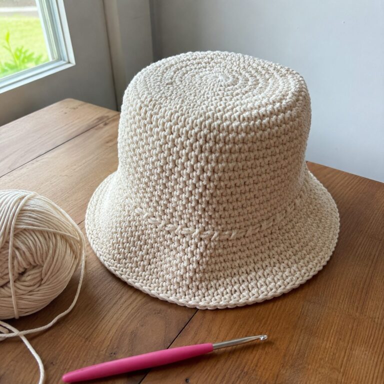 Handmade cream cotton crochet bucket hat with a soft brim sitting on a wooden table beside a ball of yarn and a pink crochet hook, natural window light