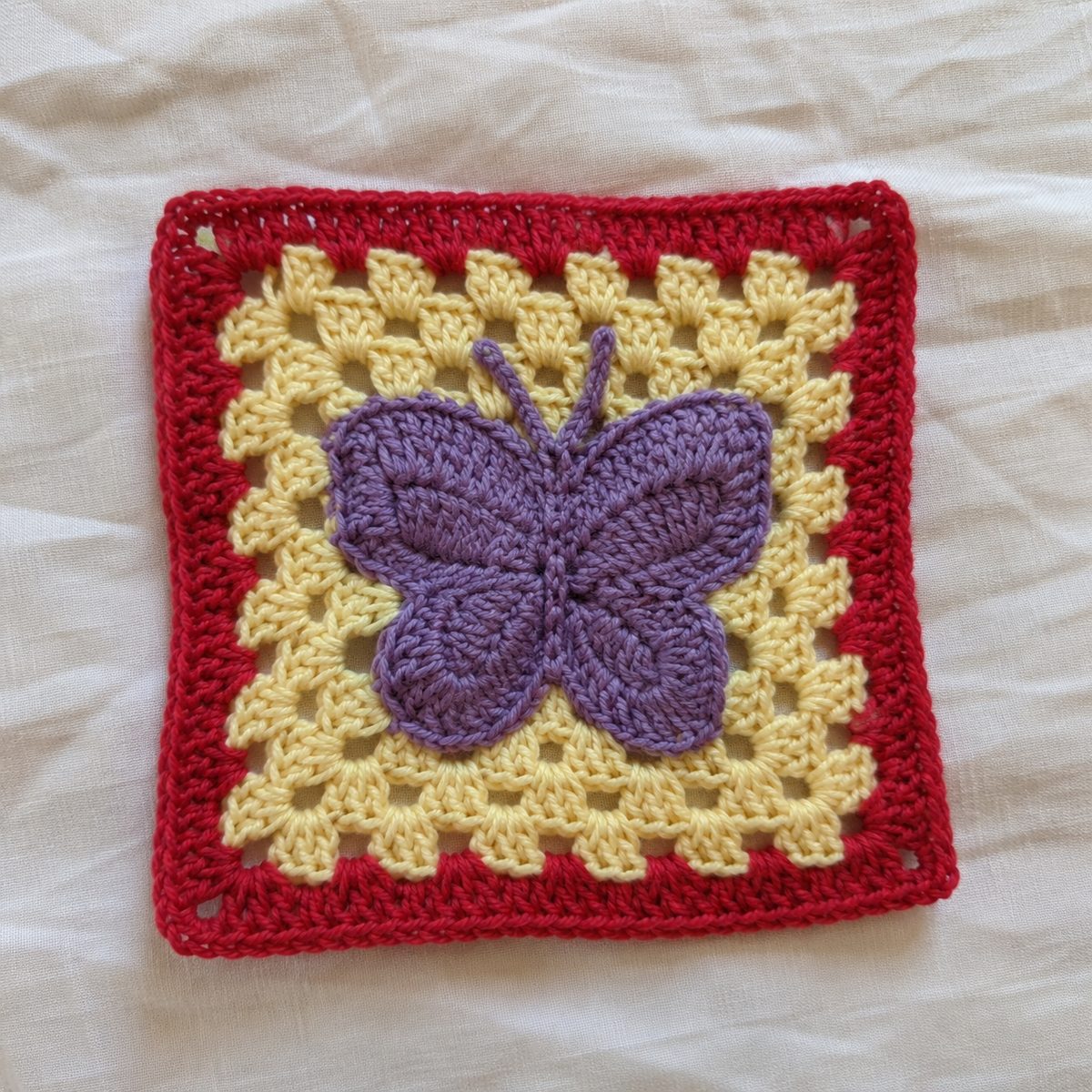 Handmade crochet granny square with a purple butterfly motif on a pale yellow background with red border, on a cream linen cloth