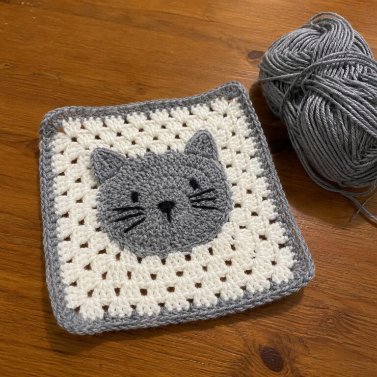 Handmade crochet granny square with a grey cat face motif with embroidered whiskers on a cream background with grey border, next to a ball of grey yarn