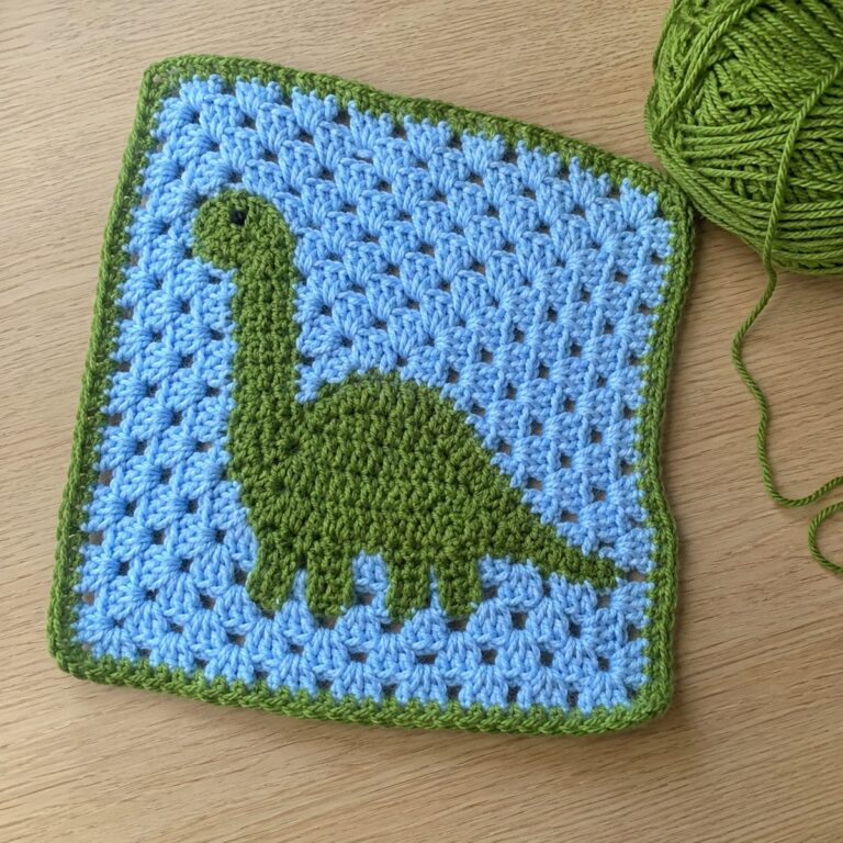 Handmade crochet granny square with a green brontosaurus dinosaur motif on a sky-blue background with green border, next to a ball of green yarn