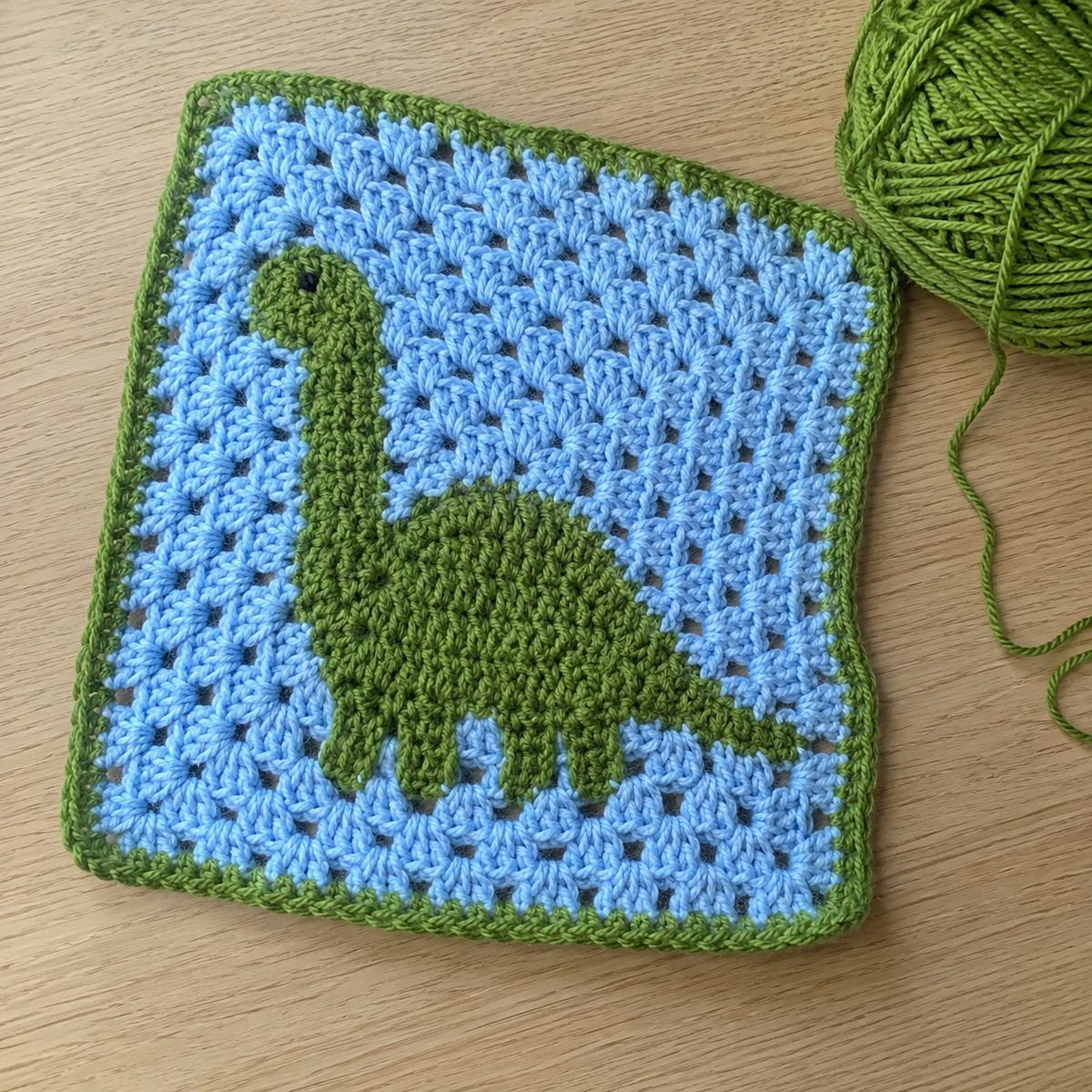 Handmade crochet granny square with a green brontosaurus dinosaur motif on a sky-blue background with green border, next to a ball of green yarn