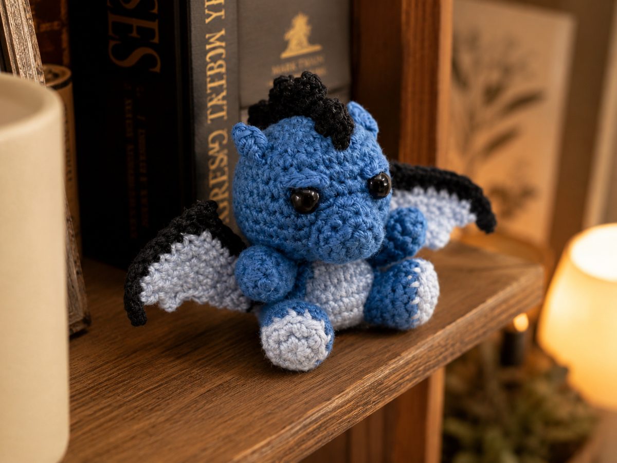 Finished amigurumi dragon sitting on a bookshelf, showing the black head crest, ears and belly patch