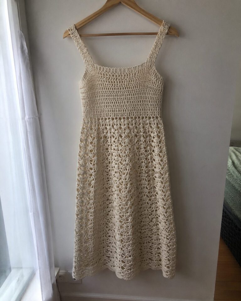 Handmade mid-length cream crochet dress with square straps and open lace shell stitch skirt, hanging on a wooden hanger against a plain wall by a real window