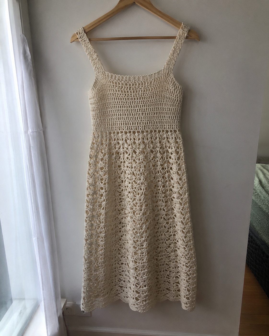 Handmade mid-length cream crochet dress with square straps and open lace shell stitch skirt, hanging on a wooden hanger against a plain wall by a real window