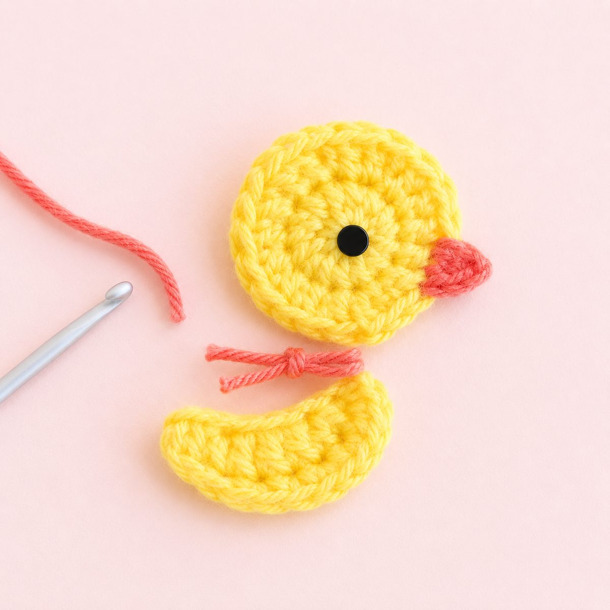 Yellow crochet duck applique in progress with a coral beak, a separate wing piece and a silver hook on a pink background