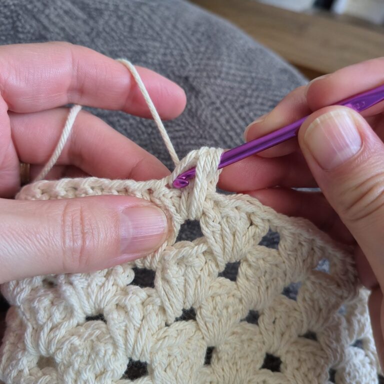 Close-up of hands working the granny spike stitch: a purple crochet hook mid-stitch making a tall spike stitch through the row below in cream cotton