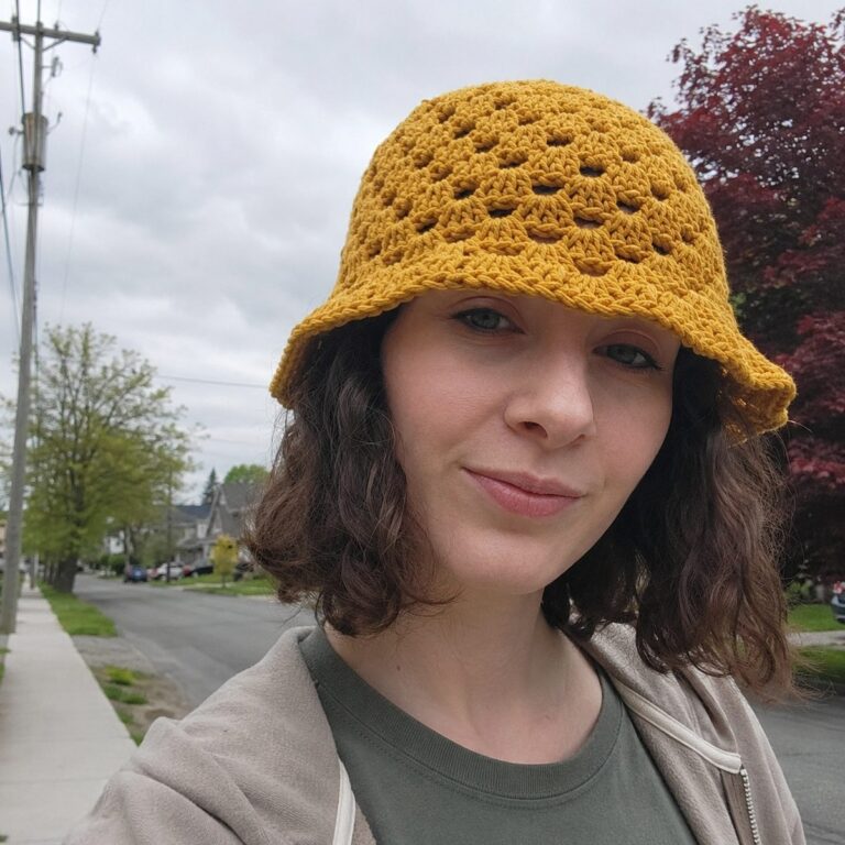 Woman wearing a handmade mustard-yellow granny stitch crochet bucket hat outdoors on an overcast day, tree-lined street in background