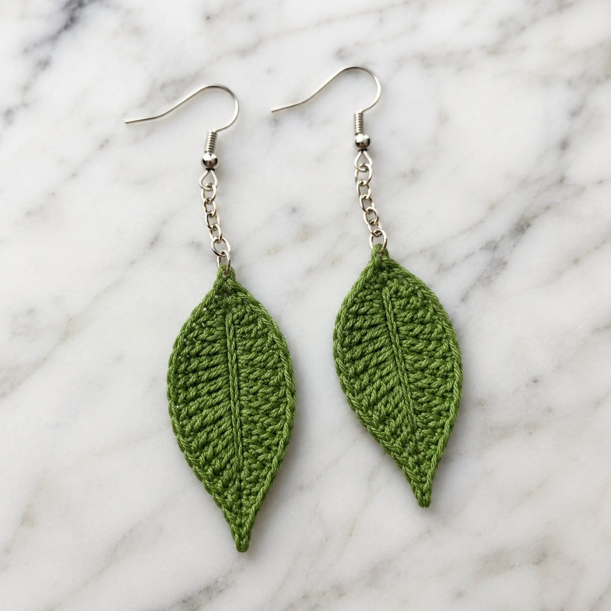 Green crochet leaf drop earrings with center veins hanging from short chains on marble