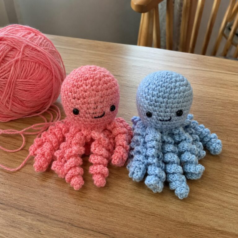 Two handmade amigurumi crochet octopuses in coral pink and pale blue with curly tentacles sitting side by side on a wooden table beside a ball of pink yarn