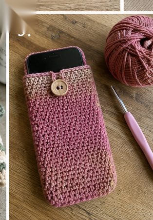 Dusty rose crochet phone case in tight single crochet with a small wooden button flap holding a smartphone, on a wooden table beside a ball of yarn and pink hook