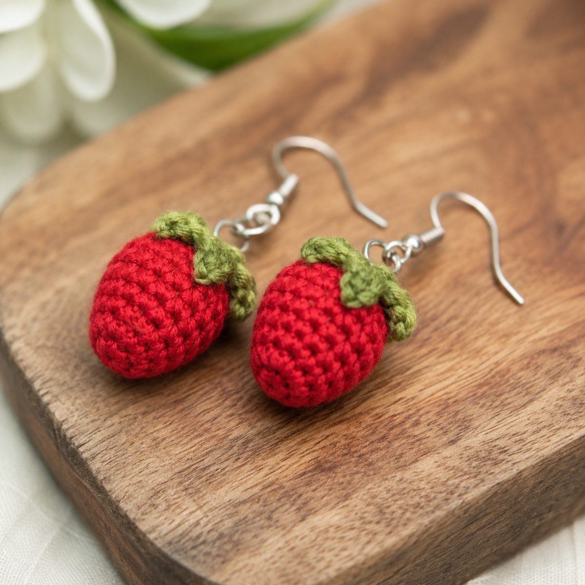 Tiny crochet strawberry earrings with green leafy tops and yellow seed stitches on a wooden board