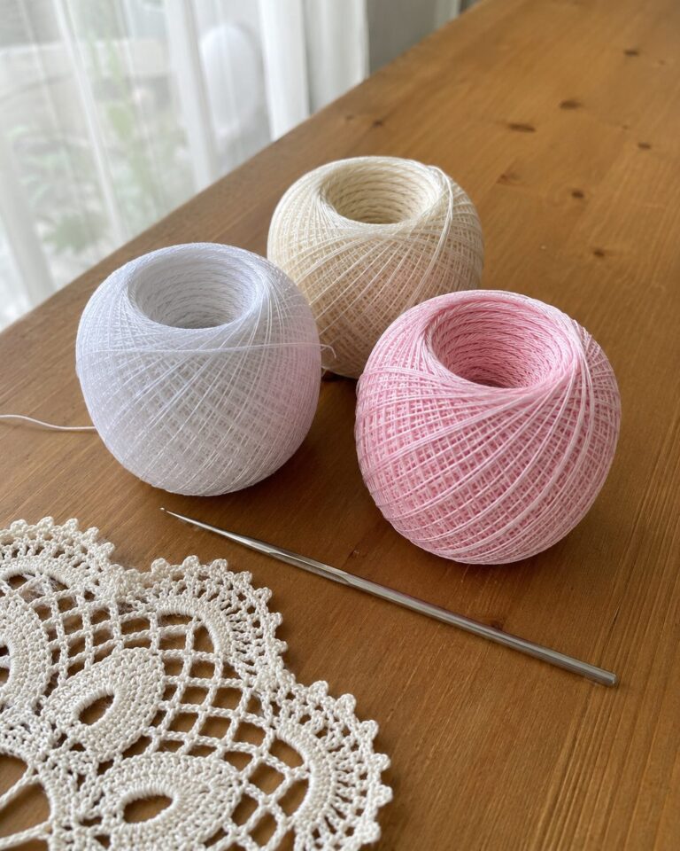 Three balls of fine crochet thread in white, ecru and pale pink on a wooden table beside a small steel crochet hook and a lace doily, natural daylight showing thread finer than yarn