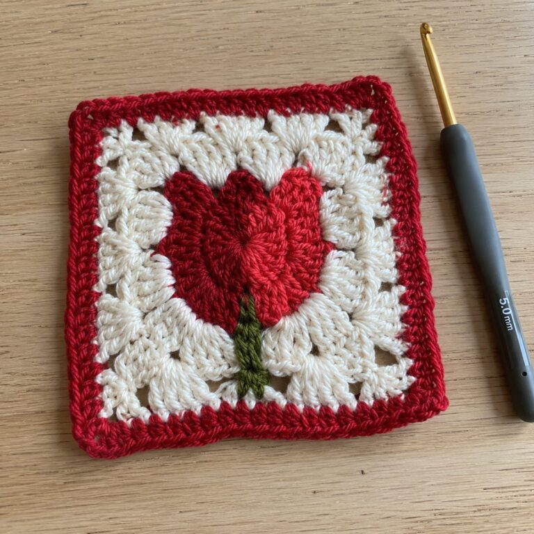 Handmade crochet granny square with a red tulip motif and green stem on a cream background with red border, on a light wood table with a crochet hook