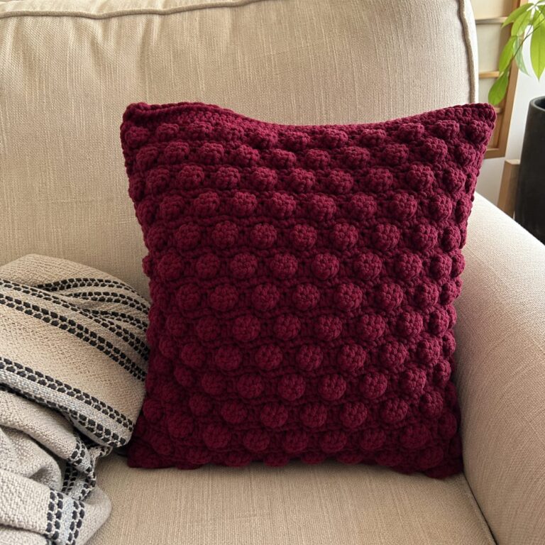Handmade burgundy crochet velvet-yarn cushion cover with a bobble stitch texture sitting on a beige linen sofa beside a striped throw