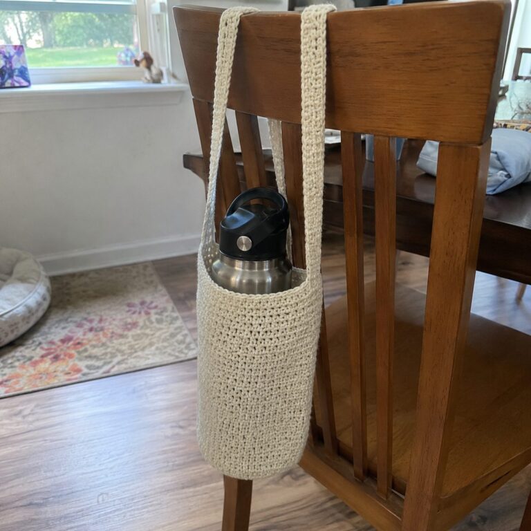 Handmade cream crochet water bottle holder with a long shoulder strap hanging on a wooden chair with a metal water bottle inside, real living room in the background