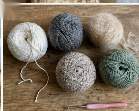 Five balls of crochet yarn in different fibers - white cotton, grey wool, fuzzy mohair, a beige blend and sage green - on a wooden table with a pink crochet hook