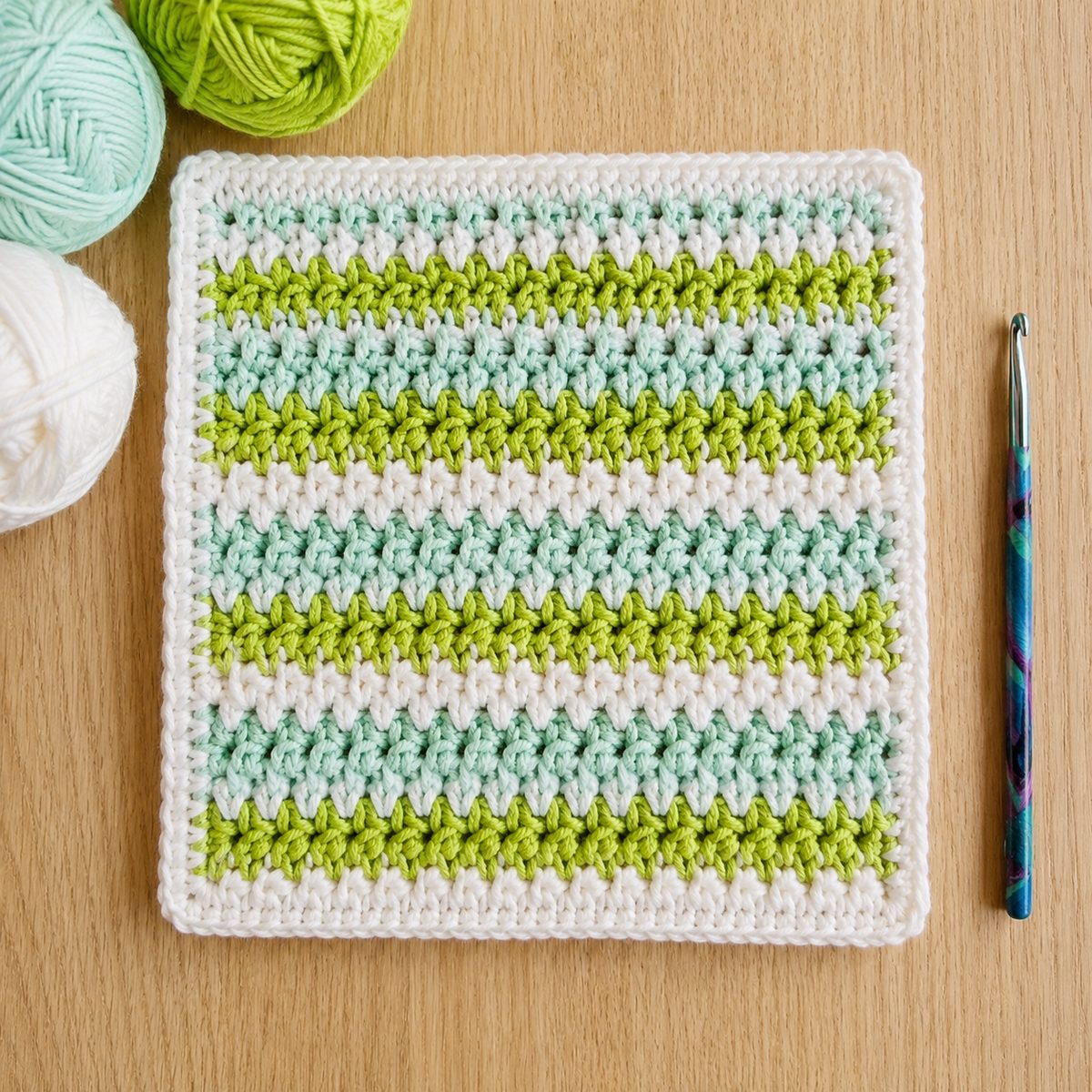 Striped crochet washcloth in white, mint and green cotton with a white border and a teal hook beside it