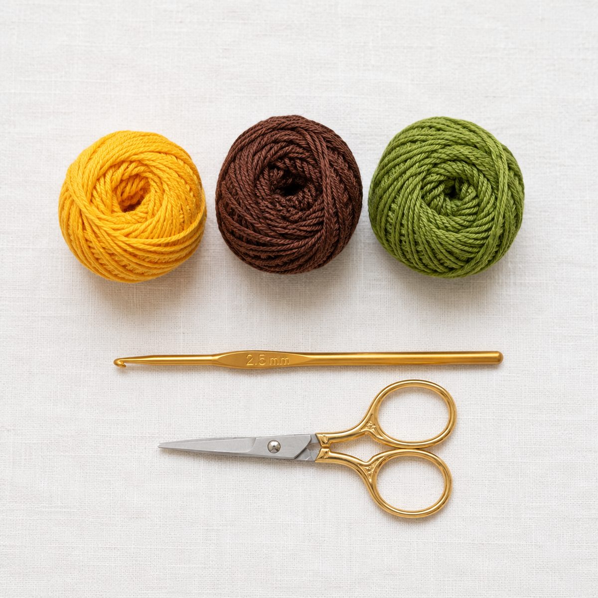 Yellow, brown and green cotton yarn balls with a 2.5 mm hook and gold scissors on white linen
