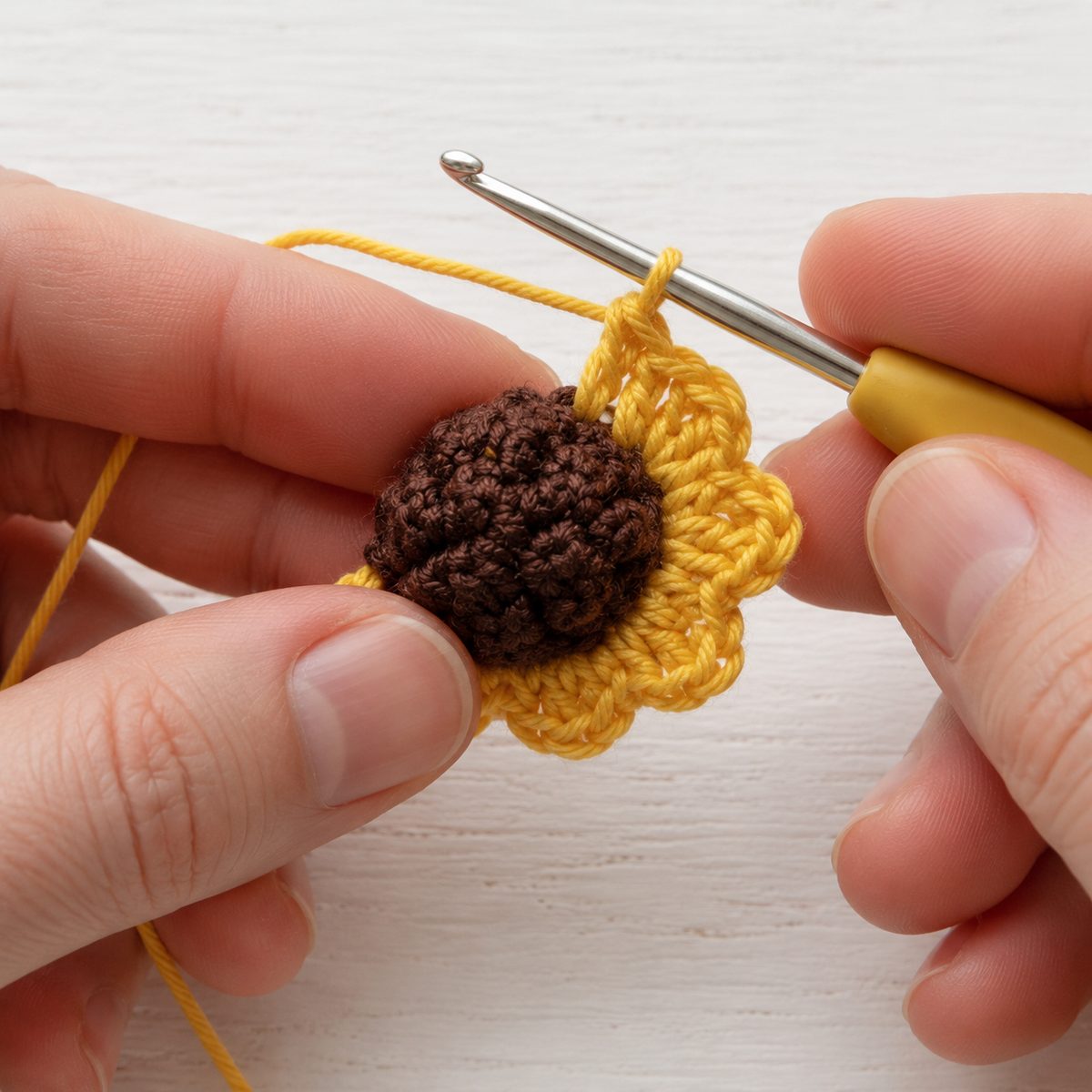 Yellow shell petals being crocheted around a brown popcorn sunflower center, hook in the working loop