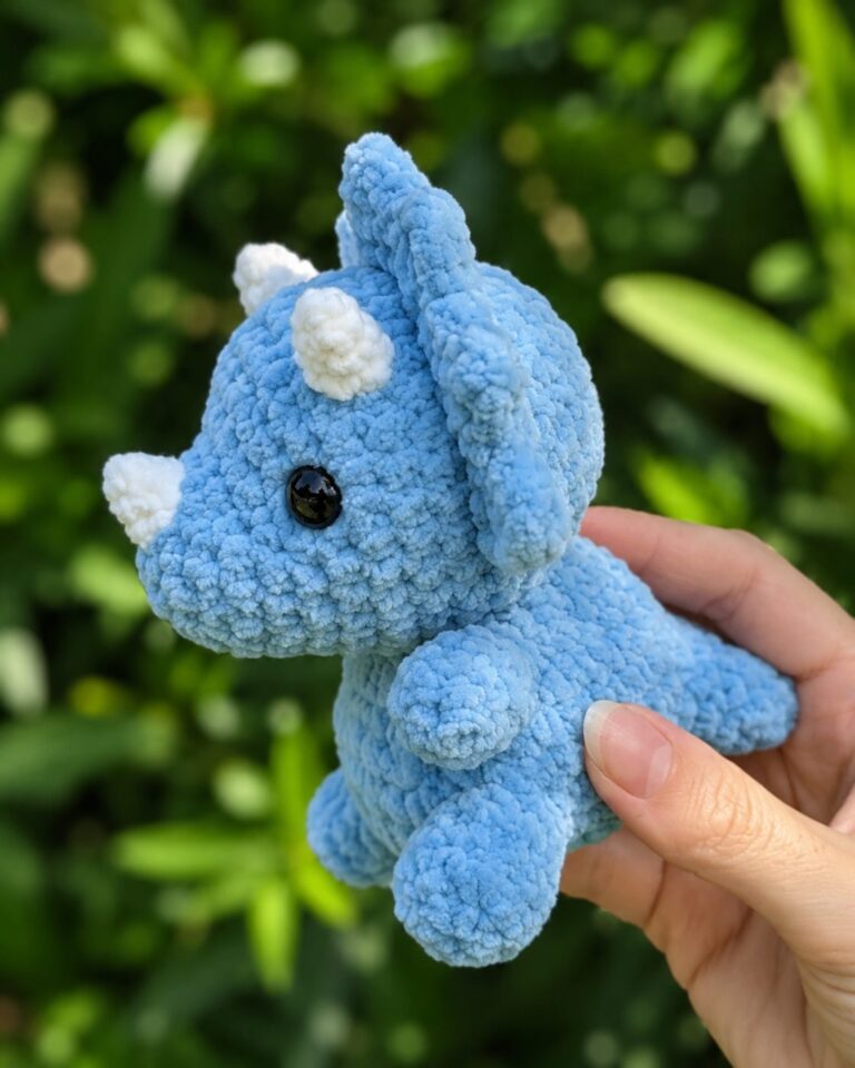 Triceratops crochet pattern finished, blue plush amigurumi dinosaur with white horns and frill held up outdoors