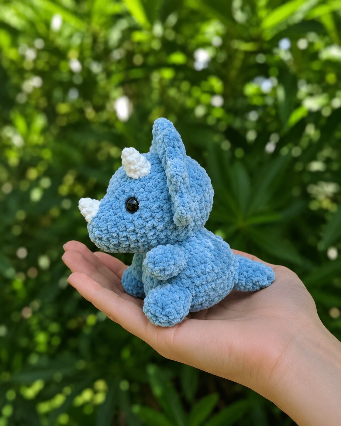 Side view of the no-sew crochet triceratops sitting in an open palm, chunky blue chenille yarn
