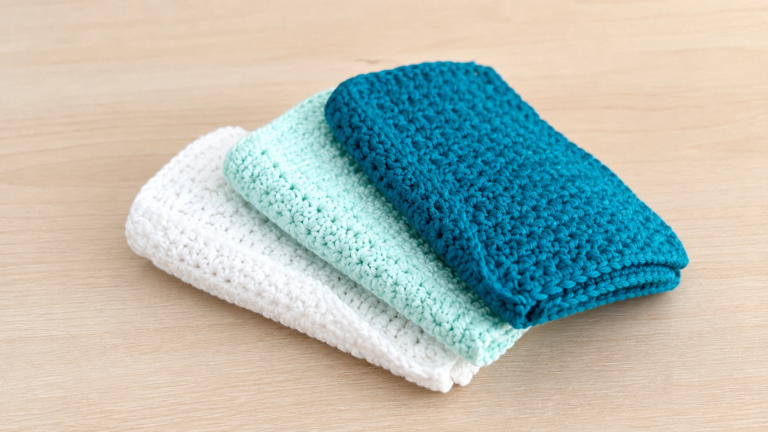 Three folded crocheted rectangular cloths in white pale mint and teal on a wooden surface