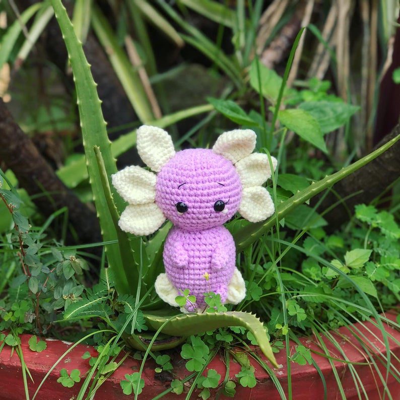 Purple crochet amigurumi axolotl with cream gills sitting among plants