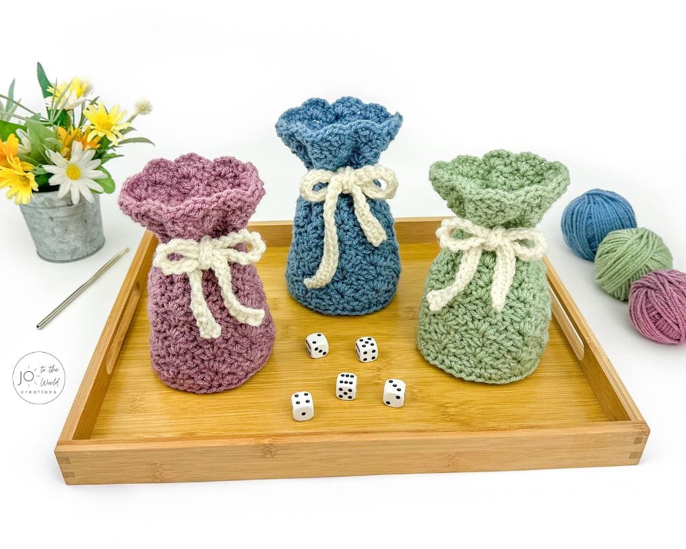 Three crochet drawstring bags in mauve, teal, and sage with white drawstring bows on a wooden tray
