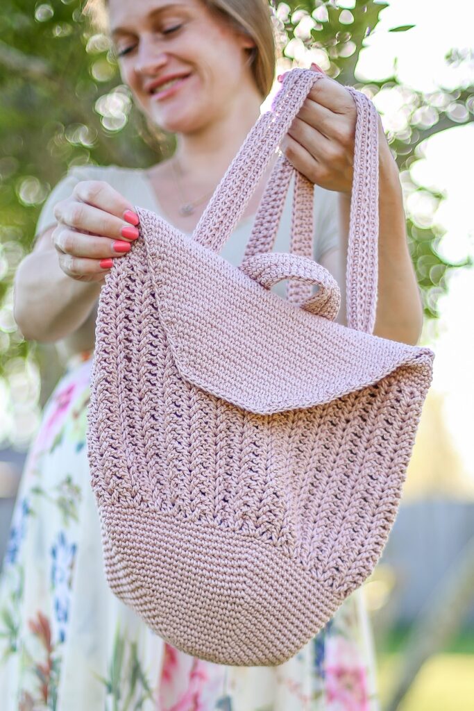 Pink crochet drawstring backpack with flap held by a woman outdoors